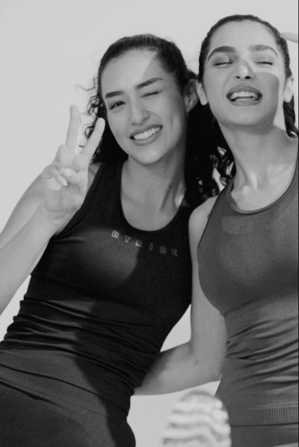 Two women posing together, one making a peace sign, against a plain background.